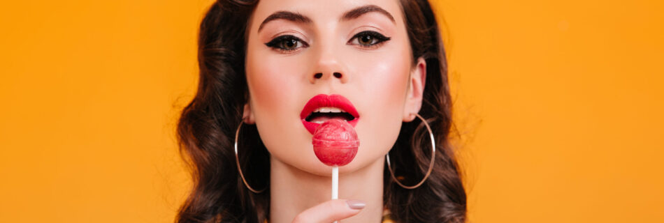 closeup view woman licking lollipop pinup girl with curly hairstyle holding candy yellow background scaled