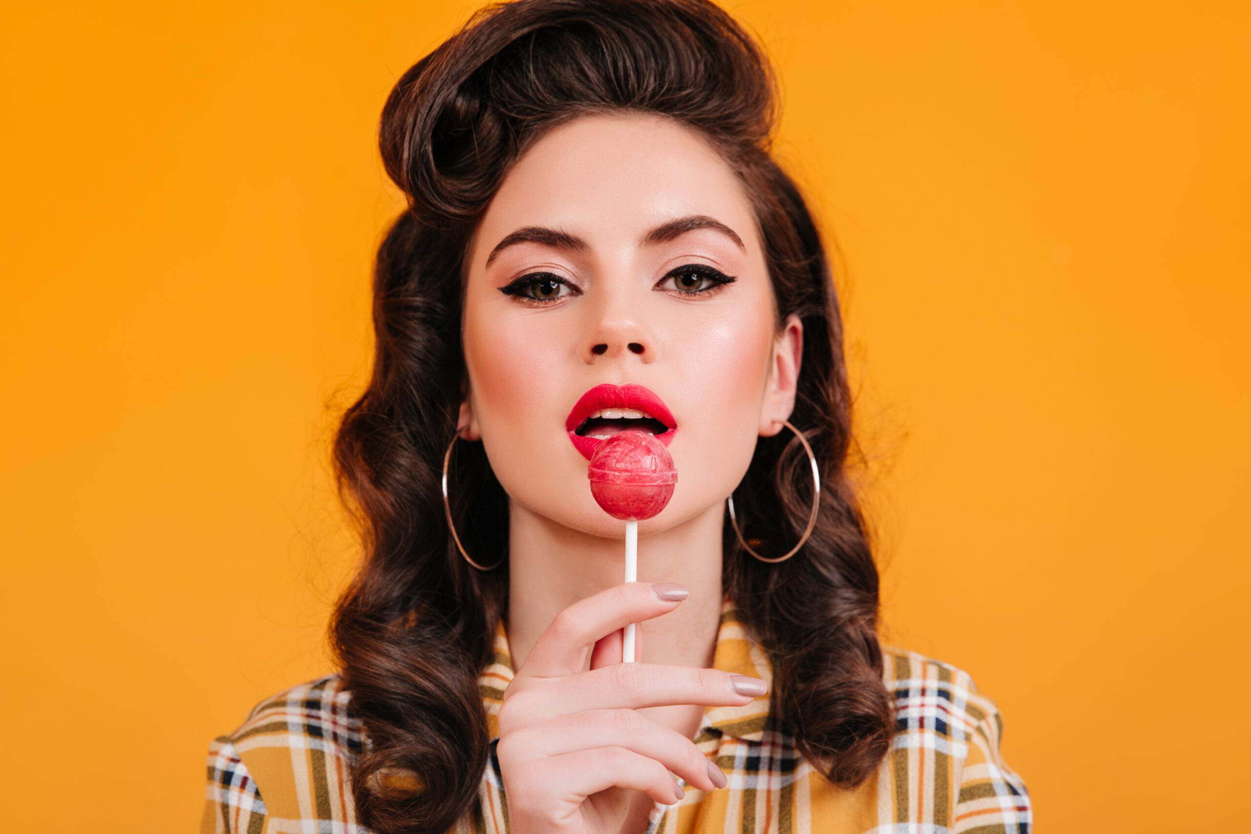 closeup view woman licking lollipop pinup girl with curly hairstyle holding candy yellow background scaled