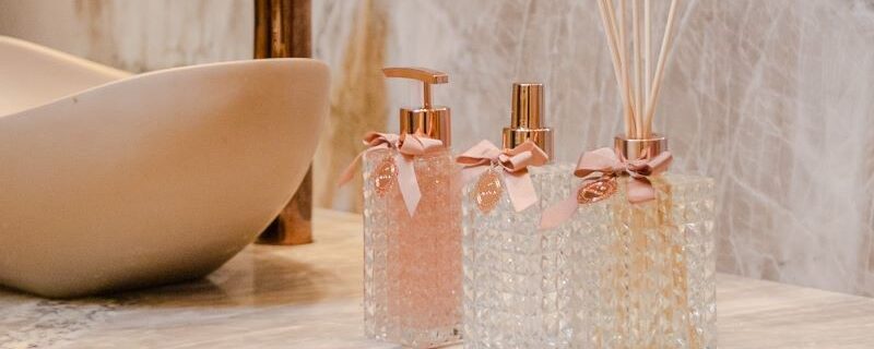 Imagem de uma pia de banheiro elegante com uma torneira de cobre e uma bancada de mármore. Sobre a bancada, há dois aromatizantes de ambiente em frascos de vidro com design cristalino, decorados com laços cor-de-rosa e varetas de difusão, ao lado de um dispensador de sabonete líquido com detalhes em cobre.